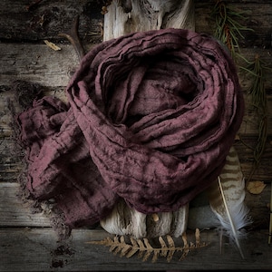 Può includere: Sciarpa bordeaux con bordo sfilacciato drappeggiata su legno. Il tessuto strutturato è disposto a cerchio. Una piuma e foglie secche completano la composizione, creando un'atmosfera rustica.