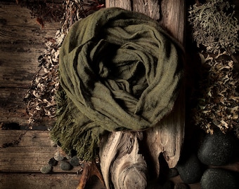 May include: An olive green scarf with fringe detail is draped over a piece of driftwood. The scarf is surrounded by dried plants, small stones, and a deer antler on a rustic wooden surface. The scarf is a solid color.