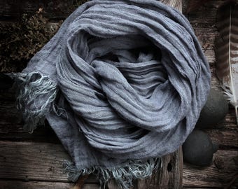 May include: A gray scarf with fringed edges is artfully arranged on a rustic wooden surface. The scarf's fabric has a textured appearance, and the overall aesthetic is natural and earthy. The scarf is a versatile accessory.