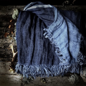 Puede incluir: Una bufanda azul oscuro con una sección azul claro y una raya blanca. La bufanda tiene un borde con flecos y está hecha de un material suave y tejido. Se encuentra sobre una superficie de madera.