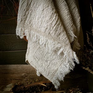 May include: A cream-colored, fringed linen throw blanket. The blanket has a textured weave and a rustic, bohemian aesthetic. The fringe detail adds a touch of elegance. The blanket is draped over a wooden surface, creating a cozy and inviting feel.