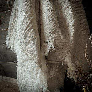 May include: A cream-colored, textured throw blanket with a fringed edge. The blanket is draped over a wooden surface, with the fringe hanging down. The fabric appears to be woven, with a slightly rough texture. The overall aesthetic is rustic and cozy.