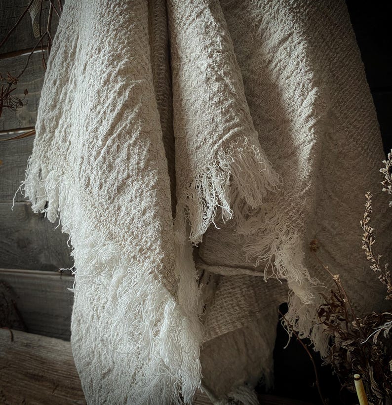 May include: A cream-colored, textured throw blanket with a fringed edge. The blanket is draped over a wooden surface, with the fringe hanging down. The fabric appears to be woven, with a slightly rough texture. The overall aesthetic is rustic and cozy.