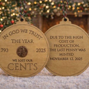 Puede incluir: Adorno navideño de madera con el texto "IN GOD WE TRUST" y "WE LOST OUR CENTS". El adorno presenta una moneda de un centavo y los años 1793 y 2025. El segundo adorno dice "DUE TO THE HIGH COST OF PRODUCTION, THE LAST PENNY WAS MINTED NOVEMBER 12, 2025."