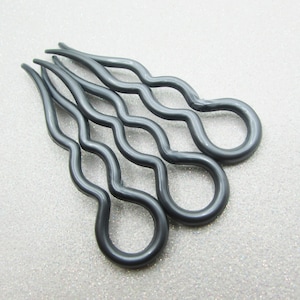 May include: Three dark gray hair pins with a wavy design. Each pin has a loop at the bottom and a series of curves leading to a three-pronged top. The pins are arranged on a textured, light gray surface.