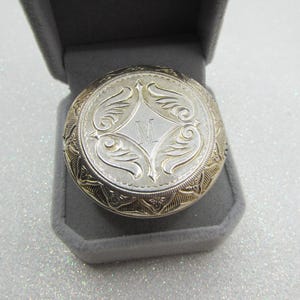 May include: A silver-toned, round compact mirror with an engraved floral design and the letter "M" in the center. The mirror is in a gray velvet box, with a gold-toned border. The compact is a vintage style.