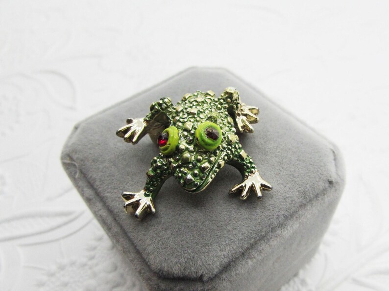 May include: A green frog brooch with a textured body, gold-colored feet, and bright green eyes. The frog has red accents and is set on a gray velvet surface. This whimsical accessory adds a touch of nature-inspired charm.