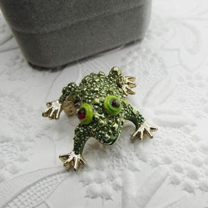 May include: A green frog brooch with gold-toned accents. The frog has textured skin, large green eyes with red centers, and gold feet. The brooch is set against a white, patterned surface, with a gray box in the background.