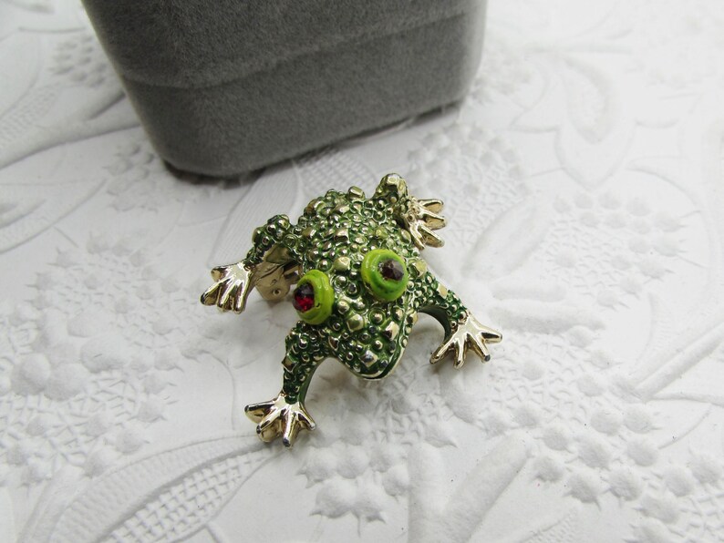 May include: A green frog brooch with gold-toned accents. The frog has textured skin, large green eyes with red centers, and gold feet. The brooch is set against a white, patterned surface, with a gray box in the background.