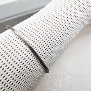 May include: A silver-toned, minimalist bangle bracelet. The bracelet has a simple, slightly twisted design. The background is a white, textured fabric, and a gray box is partially visible.