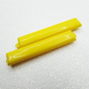 May include: Two bright yellow plastic pieces, rectangular in shape, are arranged diagonally against a white, speckled background. The pieces have a slightly rounded top and a flat bottom. The ends are also rounded.