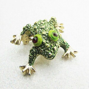 May include: A whimsical frog brooch with a textured, green enamel body and gold-tone accents. The frog has bright green eyes with red pupils and outstretched gold-tone feet. This unique pin adds a touch of nature-inspired charm to any outfit.