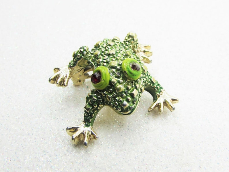 May include: A whimsical frog brooch with a textured, green enamel body and gold-tone accents. The frog has bright green eyes with red pupils and outstretched gold-tone feet. This unique pin adds a touch of nature-inspired charm to any outfit.