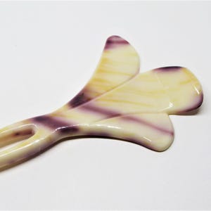 May include: A white and purple hair comb with a flower design. The comb has two prongs and a decorative flower design with a swirl pattern.