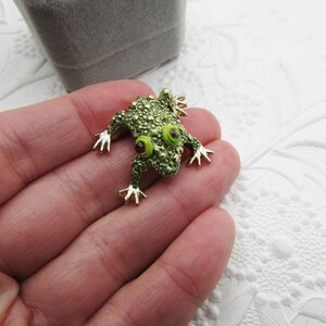 May include: A green frog brooch with gold-colored accents. The frog has large, round eyes with red centers and is covered in small, textured details. The brooch is set against a white, textured background.