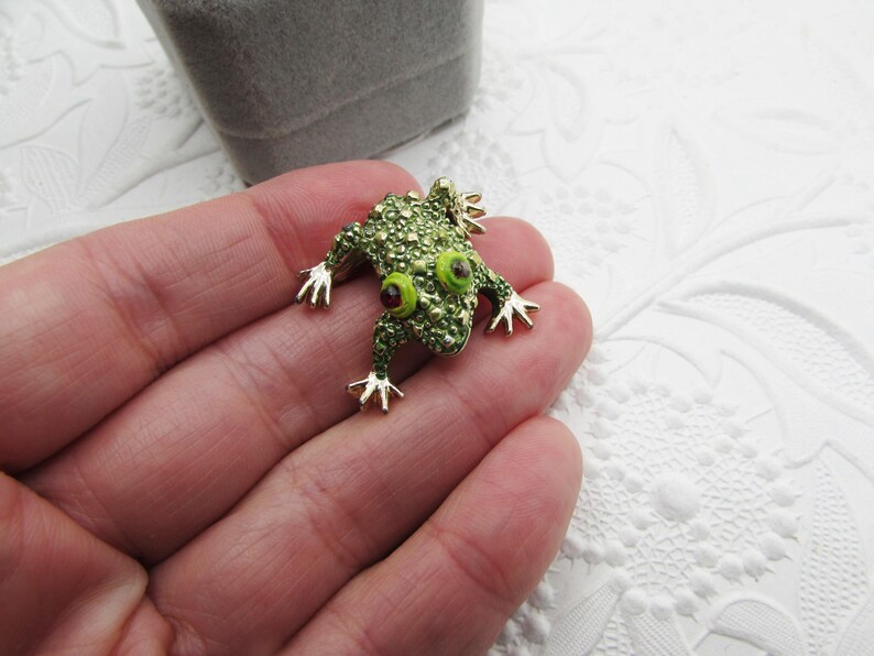 May include: A green frog brooch with gold-colored accents. The frog has large, round eyes with red centers and is covered in small, textured details. The brooch is set against a white, textured background.