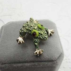May include: A detailed frog brooch with a textured, green body and gold-colored feet. The frog has large, round eyes with green rims and red centers. The brooch is set on a gray velvet surface.