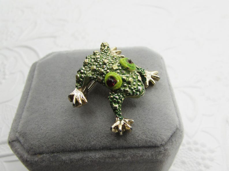 May include: A detailed frog brooch with a textured, green body and gold-colored feet. The frog has large, round eyes with green rims and red centers. The brooch is set on a gray velvet surface.