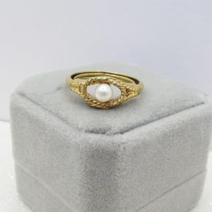 May include: A gold ring with a pearl center. The ring features a textured, rope-like design around the pearl, creating an eye-shaped setting. The ring is displayed on a gray jewelry box.