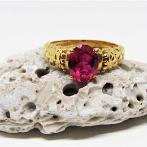 May include: A gold ring with a large, oval, ruby-colored gemstone. The ring is set on a textured, off-white stone. The ring's band has a detailed, ornate design. The gemstone is the focal point, catching the light.