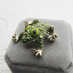 May include: A green frog brooch with gold-colored accents. The frog has textured skin, large green eyes with red centers, and gold-colored feet. The brooch is set on a gray surface.