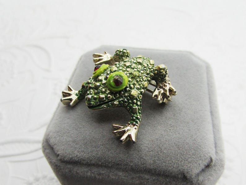 May include: A green frog brooch with gold-colored accents. The frog has textured skin, large green eyes with red centers, and gold-colored feet. The brooch is set on a gray surface.