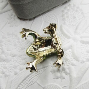 May include: A gold-toned frog brooch with green accents. The frog is in a sitting position with its legs extended. The brooch has a pin on the back for attachment. The background is a white, textured surface.
