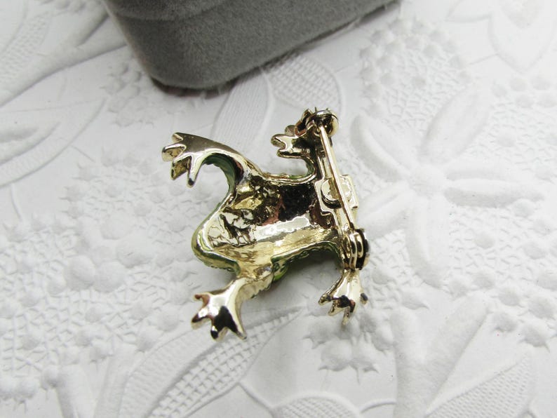 May include: A gold-toned frog brooch with green accents. The frog is in a sitting position with its legs extended. The brooch has a pin on the back for attachment. The background is a white, textured surface.