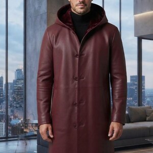 Peut inclure: Un long manteau en cuir bordeaux doté d'une capuche avec une doublure intérieure rouge foncé, de manches longues et d'une fermeture à boutons sur le devant. Le manteau est porté sur un pull à col roulé noir, créant un aspect superposé.
