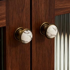 Könnte beinhalten: Nahaufnahme eines Holzschranks mit zwei Türen. Die Türen haben geriffelte Glaspaneele und sind mit Messingknöpfen mit weißen und grauen Marmor-Akzenten versehen. Der Schrank ist aus dunkelbraunem Holz gefertigt.