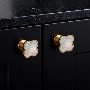 Op de afbeelding: Twee zwarte kastdeuren met goudkleurige klavervormige knoppen. De knoppen hebben een parelmoer, iriserend midden en een goudkleurige metalen rand. De kastdeuren staan tegen een zwart aanrechtblad.