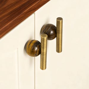 以下が含まれることがあります： 装飾的な金具が付いた2つのキャビネットドアのクローズアップ。各ドアには、真鍮色の円筒形のハンドルが1組あり、天然石の球が付いています。石の球は茶色と黄色の縞模様です。キャビネットのドアはオフホワイトです。