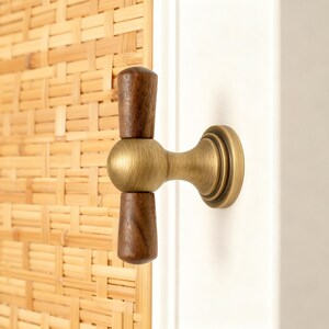 May include: A cabinet pull with a brass finish and two dark brown wooden handles. The pull is mounted on a white surface, with a woven bamboo background.