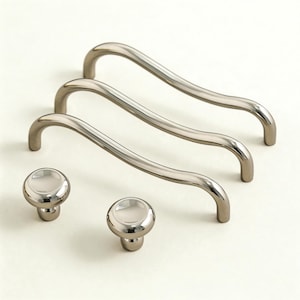 May include: A set of three curved cabinet pulls and two matching round knobs. The hardware is made of polished silver metal, reflecting light. The pulls have a gentle wave design, and the knobs have a flat, circular top.
