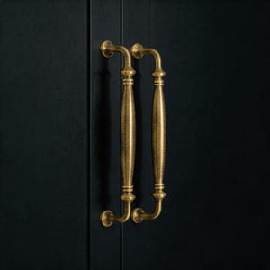 Puede incluir: Dos tiradores de armario de latón antiguo con un diseño curvo. Los tiradores están montados en una puerta de armario oscura. El latón tiene un acabado cálido y envejecido, y el diseño incluye detalles decorativos.
