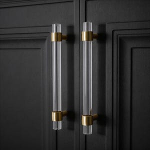 Op de afbeelding: Twee heldere acryl kastgrepen met goudkleurige metalen accenten. De grepen zijn cilindrisch en gemonteerd op een donkere houten kast. De hardware voegt een moderne touch toe.
