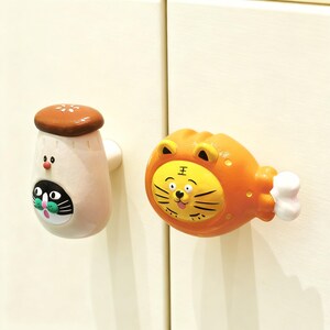 Könnte beinhalten: Zwei skurrile Schrankknöpfe. Einer ist pilzförmig mit einem Katzenkopf, der andere ist ein orangefarbenes Tigergesicht auf einer Keulenform. Beide Knöpfe haben weiße Befestigungsteile.