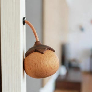 May include: A wooden door stop with a round, light-colored body and a dark brown, leaf-shaped top. A tan cord extends from the top and is inserted into a hole in the door frame. The door stop is made of wood.