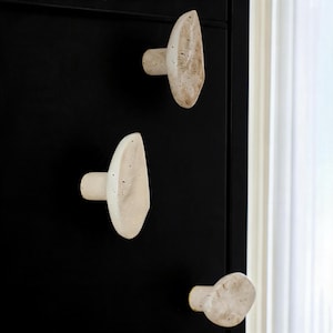 May include: Three off-white, textured cabinet knobs with an organic shape are mounted on a black cabinet door. The knobs have a cylindrical base and a flattened, asymmetrical face. The knobs are spaced vertically on the door.