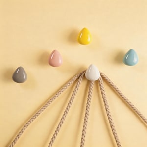 May include: A collection of five teardrop-shaped wall hooks in various colors: gray, pink, yellow, white, and blue. The hooks are mounted on a light yellow wall, with a beige rope-like structure hanging below the white hook. These hooks are designed for hanging items.