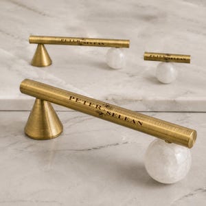 May include: Three cabinet pulls featuring a brushed brass cylindrical bar with the text PETER + SELEAN engraved on the surface. Each pull is supported by one conical brass base and one spherical white stone base, creating a mixed material design.