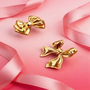 May include: Two gold-colored bow-shaped decorative objects on a pink gradient background. The bows have detailed folds and a shiny finish. Pink ribbons are draped around the bows, adding to the decorative theme.