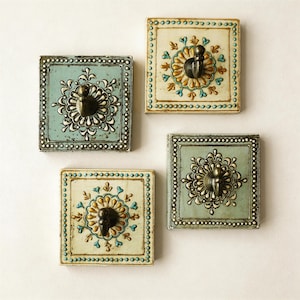 Puede incluir: Cuatro perchas de pared cuadradas decorativas. Cada percha presenta un diseño central ornamentado con un gancho de metal oscuro. Los cuadrados están pintados en tonos de azul y crema, con bordes decorativos y motivos florales.