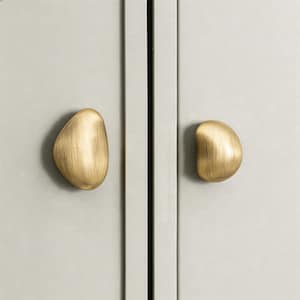 May include: Two brushed brass cabinet knobs with an organic, pebble-like shape are shown. They are mounted on a light grey cabinet door, offering a modern and minimalist aesthetic. The hardware adds a touch of elegance to the cabinet.