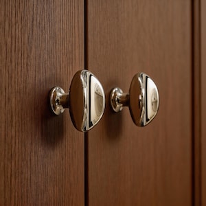 May include: Close-up of two polished chrome cabinet knobs with an oval design. The knobs are mounted on a dark brown wooden cabinet door, reflecting the surrounding environment. The hardware adds a touch of elegance.