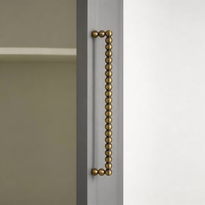 Puede incluir: Un tirador de puerta de armario con un diseño vertical. El tirador está hecho de pequeñas cuentas redondas de color latón. El tirador está fijado a una puerta de armario gris.