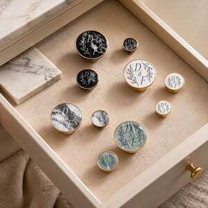 May include: A collection of round cabinet knobs in various sizes and finishes, including black, white, and sage green marble-effect surfaces. Each knob features a gold-toned metal rim and a central graphic of the letters D & F framed by leafy branches.