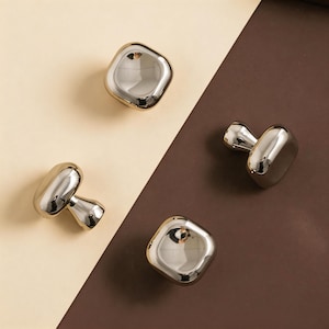 May include: Four shiny, silver cabinet knobs with a rounded, square design. Each knob has a mushroom-like shape with a polished finish. The knobs are arranged on a two-tone background of beige and brown.