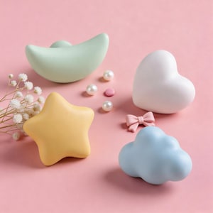 Puede incluir: Una colección de objetos decorativos en colores pastel, que incluyen una estrella amarilla, un corazón blanco, una nube azul y una luna creciente verde. También hay pequeñas perlas rosas y blancas y un lazo rosa. Los objetos están dispuestos sobre una superficie rosa.