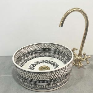 Peut inclure: Un lavabo rond en céramique blanche avec un motif géométrique noir et un robinet doré. L'évier a une bordure décorative et un drain central. Le robinet a un bec courbé et des poignées en croix.